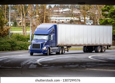 Big rig blue classic American modern semi truck with high cab for long haul routs with dry van semi trailer transporting commercial cargo driving on the turning city street going under the bridge 