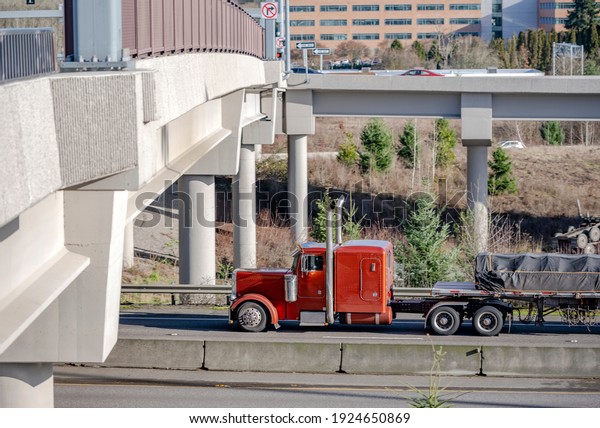 Big rig brown semi truck tractor with chrome\
vertical exhaust pipes transporting covered commercial cargo on\
flat bed semi trailer running on the divided highway road going\
under the concrete bridge