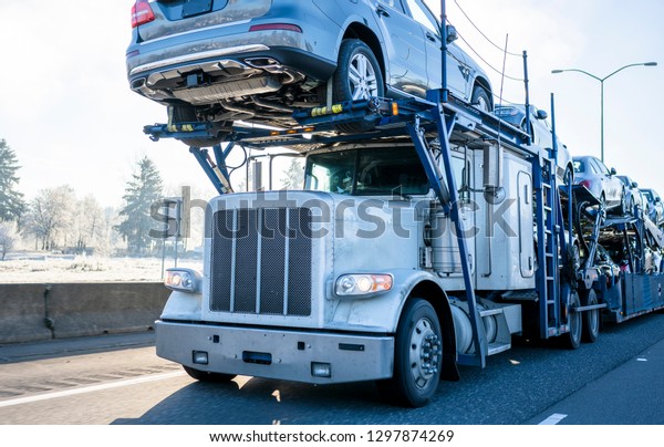 Big rig car\
hauler professional bonnet powerful semi truck transporting cars on\
two levels industrial semi trailer driving on winter frosty road\
with frost grass and trees on\
roadside