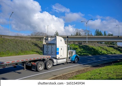 Big Rig Classic High Cab Powerful Efficient Eco-friendly Diesel Semi Truck With Storage Boxes On Back Wall Of The Cab Transporting Empty Flat Bed Semi Trailer Running On The Road With Bridge Ahead