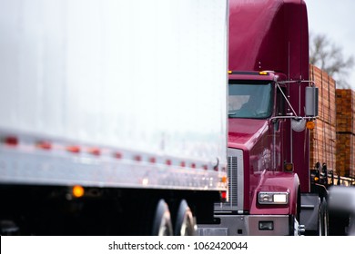 Big Rig Classic Powerful Dark Red Semi Truck Tractor Transporting Stacks Of Boards Lumber Wood On Flat Bed Semi Trailer Driving In Convoy Behind Another Dry Van Semi Truck Trailer In Rain Weather