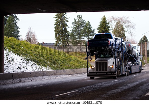 Big rig\
classic white car hauler semi truck transporting cars on special\
two-storey semi trailer going by highway under bridge in winter\
weather with snow on the shoulder\
grass