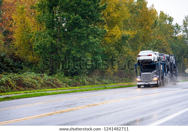 Big rig dark car hauler powerful semi truck\
transporting cars on two levels semi trailer driving with turned on\
headlight for safety on the slippery wet road in rain with autumn\
trees on the side