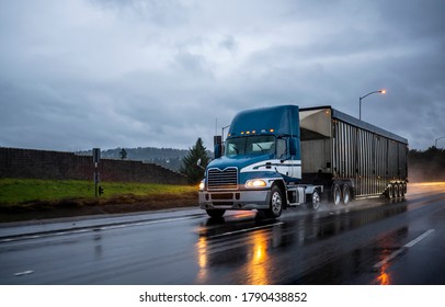 Big rig day cab bonnet blue semi truck with roof spoiler transporting commercial cargo in covered corrugated bulk semi trailer running on the evening wet glossy slippery road with raining weather