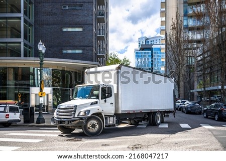 Big rig day cab white semi truck with long box trailer making local commercial delivery at urban city with multilevel residential apartments buildings turning on the city street with crossroad
