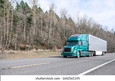 Big rig green classic powerful diesel long haul semi truck transporting frozen commercial cargo in corrugated refrigerated semi trailer running on the road with bare trees forest on the hill
