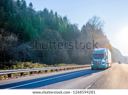 Big rig green modern bonnet American semi truck with high cab for long haul routs transporting refrigerated semi trailer driving on the straight highway in front of another traffic in sunny day