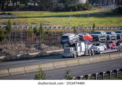 Big rig industrial grade white car hauler semi truck transporting crossover cars on the two level hydraulic modular semi trailer running on the straight divided multiline highway road at sunny day