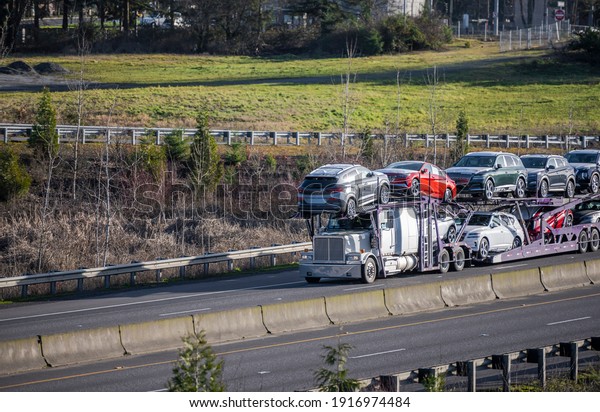 Big rig industrial grade white car hauler semi truck\
transporting crossover cars on the two level hydraulic modular semi\
trailer running on the straight divided multiline highway road at\
sunny day