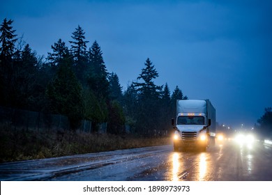 Big rig industrial white semi truck with turned on headlights transporting cargo in dry van semi trailer running on the night dark twilight wet road with reflection on surface in rain weather 