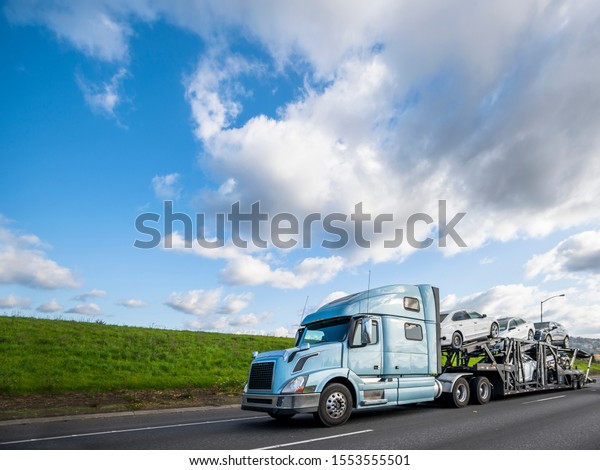 Big rig light blue long haulage car hauler semi\
truck with drivers rest compartment transporting different cars on\
the special two levels modular semi trailer driving on the flat\
road with green hill