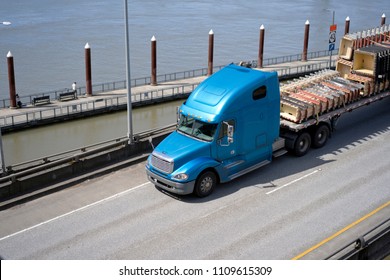 Big Rig Long Haul Blue Semi Truck With Tall Cab And Flat Bed Semi Trailer Transporting Commercial Cargo On Elevated Road Along The River With Pier Posts On The Background