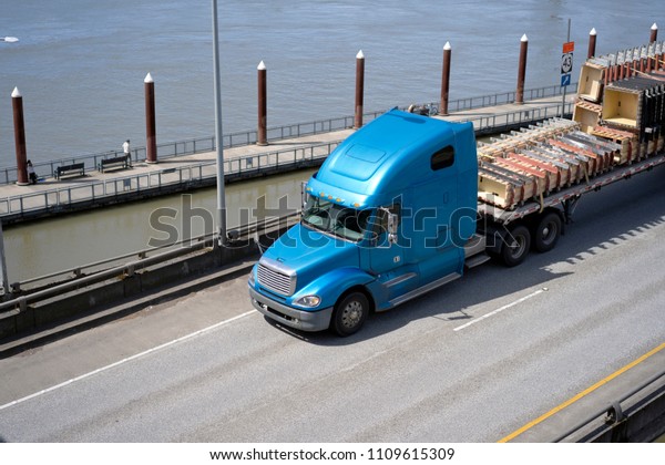 Big rig long haul\
blue semi truck with tall cab and flat bed semi trailer\
transporting commercial cargo on elevated road along the river with\
pier posts on the\
background