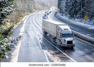 Big rig long haul gray semi truck tractor transporting commercial cargo in dry van semi trailer going on the wet slippery road with water from melting snow and winter snowy trees on the hills