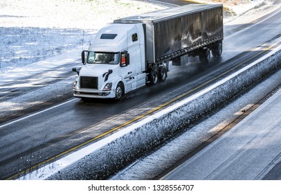 Big rig long haul gray semi truck tractor transporting commercial cargo in dry van semi trailer going on the wet slippery road with water from melting snow and winter snowy trees on the hills