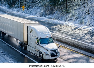 Big Rig Long Haul Gray Semi Truck Tractor Transporting Commercial Cargo In Dry Van Semi Trailer Going On The Wet Slippery Road With Water From Melting Snow And Winter Snowy Trees On The Hills