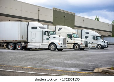 Big rig long haul industrial freight white semi truck loading commercial cargo to dry van semi trailer standing at the warehouse dock gate in row with another loaded semi trucks and semi trailers