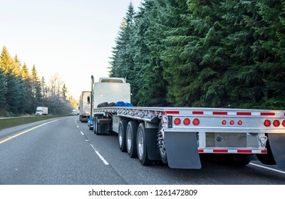 Big rig long haul powerful semi truck with empty light weight aluminum flat bed semi trailer running on the winter road in convoy with another semi trucks and evergreen trees