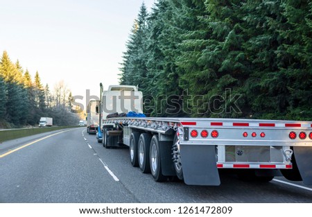 Big rig long haul powerful semi truck with empty light weight aluminum flat bed semi trailer running on the winter road in convoy with another semi trucks and evergreen trees
