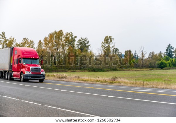 Big rig long haul\
red bonnet powerful semi truck tractor transporting refrigerator\
semi trailer with commercial cargo on the straight road with trees\
and field background