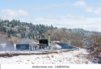 Big Rig Long Haul Semi Truck Tractor Transporting Empty Semi Trailer Going To Warehouse For Loading Cargo Running On Wet Glossy Road With Water From Melting Snow And Winter Snowy Trees On The Hill