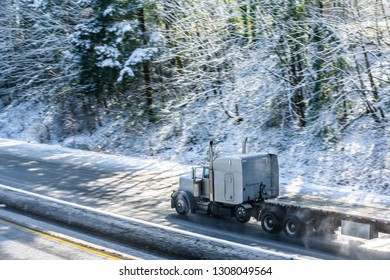 Big rig long haul semi truck tractor transporting empty semi trailer going to warehouse for loading cargo running on wet glossy road with water from melting snow and winter snowy trees on the hill