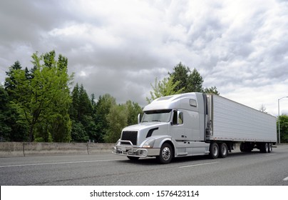 Big rig long haul semi truck with pipe grille guard transporting commercial cargo in refrigerator semi truck running on the wide highway with trees and clouds on the background