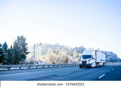 Big rig long haul white semi truck with high cab transporting refrigerator unit on reefer semi trailer driving on the winter wide multiline highway with snow and frosted trees on the hillside