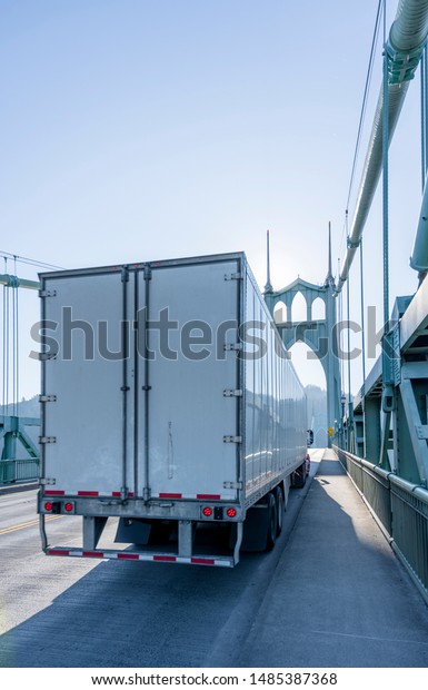 Big rig long haul white semi\
truck transports cargo in semi trailer running on the St Johns\
bridge with arch gothic support tower and rope stretching in\
sunshine