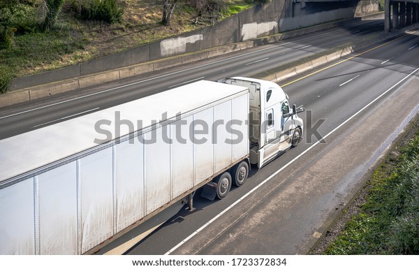 Big rig long haul white semi truck with high cab
configuration for improve aerodynamics transporting cargo in loaded
dry van semi trailer running on the road under the bridge across
divided highway