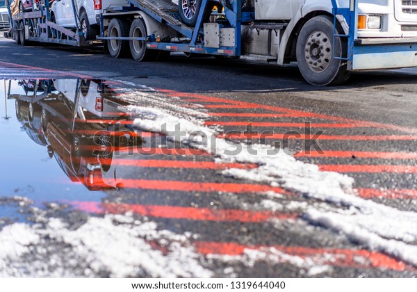 Big rig long hauler commercial freight classic\
car hauler semi truck transporting cars on modular hydraulic semi\
trailer and reflecting in a puddle of melted snow on the surface\
with road markers