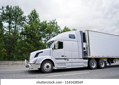 Big Rig Modern White Long Haul Semi Truck With Protection Grille Guard Refrigerator Semi Trailer Transporting Perishable And Frozen Foods On The Wide Highway Road With Trees On The Side