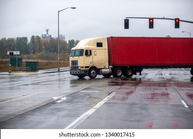 Big Rig Old Cab Over American Classic Long Haul Semi Truck With Container On Flat Bed Semi Trailer Turning On Traffic Light Crossroad Intersection Wet Raining Road With Reflection Going To Warehouse