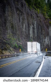 Big Rig Powerful Blue Semi Truck With Pipes Grill Guard Transporting Commercial Cargo In Semi Trailer Running On The Winding Wet Road With Rain Dust With Rock Cliff On The Side In Columbia Gorge