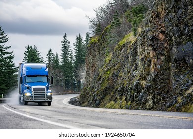 Big rig powerful blue semi truck with pipes grill guard transporting commercial cargo in semi trailer running on the winding wet road with rain dust with rock cliff on the side in Columbia Gorge