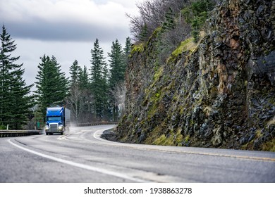 Big Rig Powerful Blue Semi Truck With Pipes Grill Guard Transporting Commercial Cargo In Semi Trailer Running On The Winding Wet Road With Rain Dust With Rock Cliff On The Side In Columbia Gorge