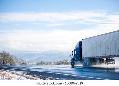Big rig pro long haul blue semi truck tractor transporting commercial cargo in refrigerator semi trailer going on the wet glossy road with water from melting snow and winter snowy trees on the side