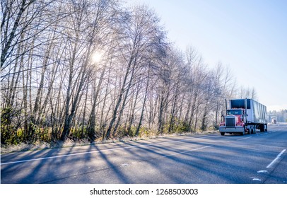 Big rig red classic day cab stylish American semi truck with high exhaust pipes and covered dry van semi trailer with front wall spoiler going on the winter road with frosted trees in sunshine