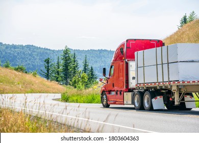 Big rig red classic long haul semi truck with flat bed semi trailer transporting tightened packed industrial lumber boards running on the turning winding road with hills and trees in sunny day