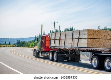 Big rig red classic powerful long haul semi truck with flat bed semi trailer transporting tightened industrial lumber boards running on the turning winding road with hills and trees in sunny day