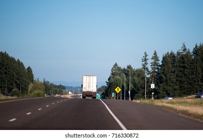 A Big Rig Semi Truck Tractor With A Dry Van Semi Trailer Moves Towards The Horizon Along A Wide Marked Multi-lane Highway Framed Along The Sides Of A Rows Of Ever-green Trees