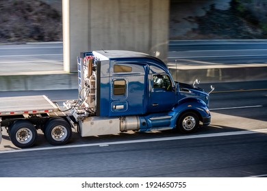 Big rig shiny blue semi truck tractor with long cab and auxiliary equipment on the back wall transporting flat bed semi trailer driving on the highway road under the concrete bridge - Shutterstock ID 1924650755