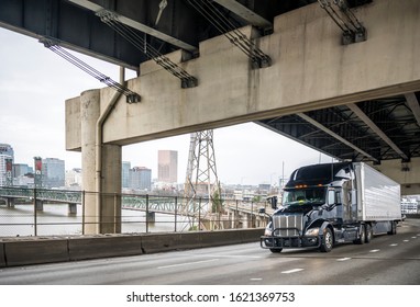 Big rig stylish black long haul semi truck with pipes grille guard transporting frozen commercial cargo in refrigerator semi trailer running on the lower level of two levels bridge across the river - Shutterstock ID 1621369753