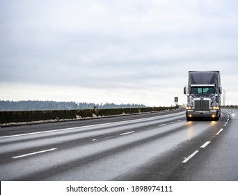 Big rig stylish industrial dark gray semi truck with turned on headlights transporting cargo in dry van semi trailer running on the twilight wet road with light reflection surface in rain weather 