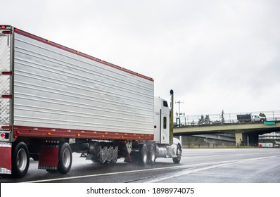 Big rig stylish industrial white semi truck with turned on headlights transporting cargo in refrigerator semi trailer running on the twilight wet road with light reflection surface in rain weather 