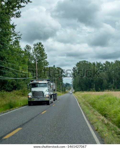 A big rig truck\
hauls its freight along a two-lane country road that cuts through\
farmlands and woodlands.  The sky is cloudy. In Washington State in\
the Pacific Northwest