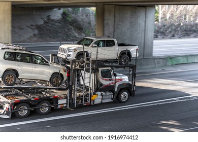 Big rig white car hauler semi truck transporting cars on the two level hydraulic modular semi trailer running on the wide divided highway with transportation bridge and overpass road intersection