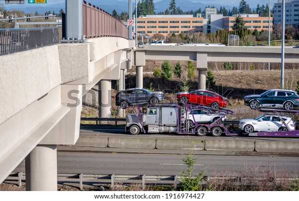 Big rig white car hauler semi truck\
transporting cars on the two level hydraulic modular semi trailer\
running on the wide divided highway with transportation bridge and\
overpass road intersection