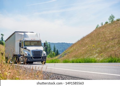 Big Rig White Day Cab Semi Truck With Roof Spoiler Transporting Huge Covered Bulk Semi Trailer Moving Uphill On The Turning Winding Road With Yellow Grass And Green Trees