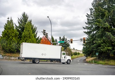 Big Rig White Day Cab Commercial Semi Truck For Local Deliveries With Long Box Trailer Turning On Freeway Entrance From City Street Road With Traffic Lights And Road Sign Arrows And Trees On The Sides